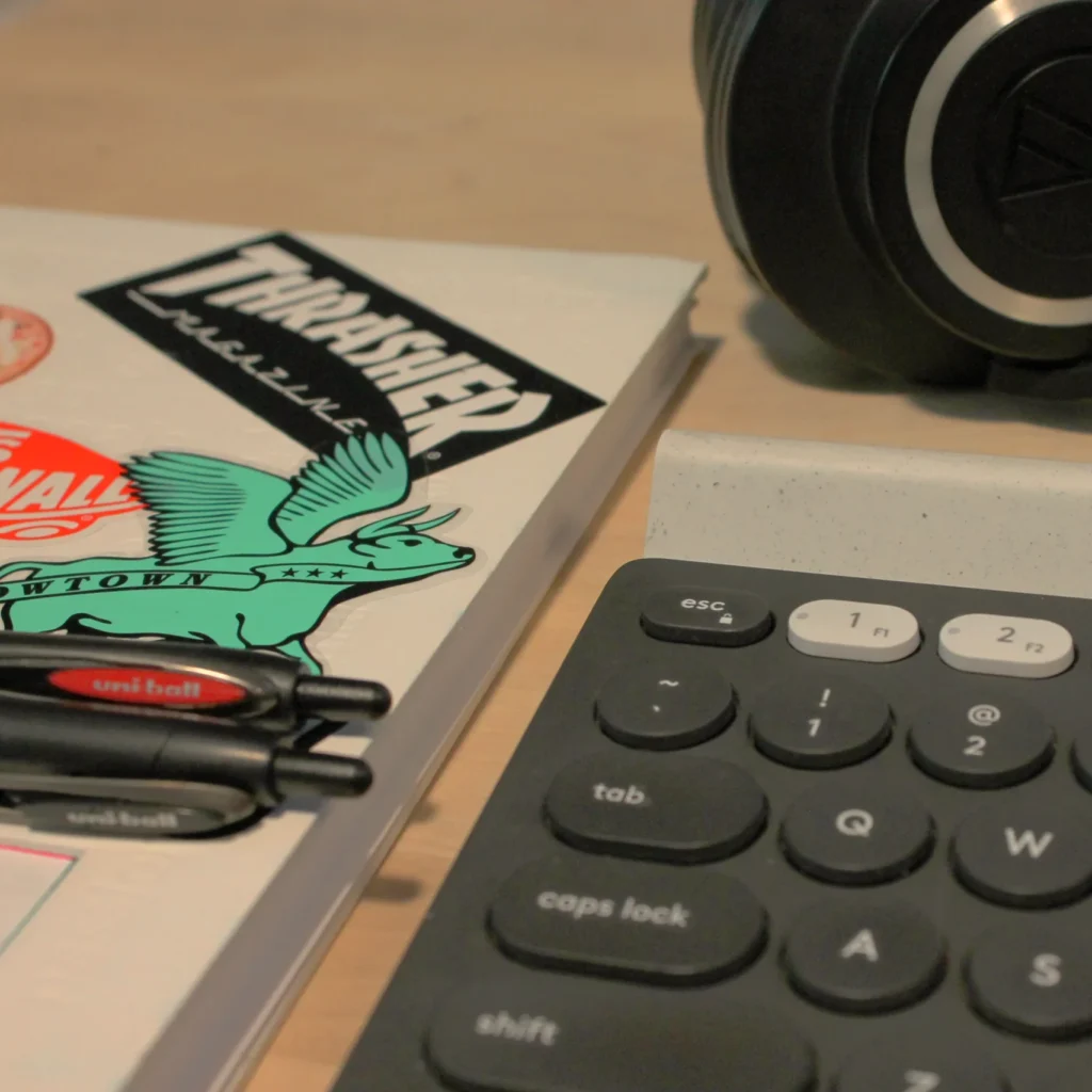 A notebook with pens on it, next to a computer keyboard and headphones on top of a desk.