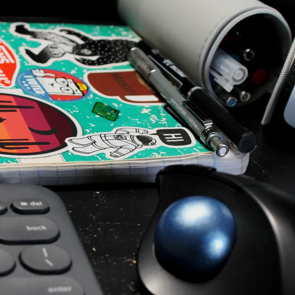A pen and pencil, sitting on a notebook next to a pencil holder, keyboard, and mouse.
